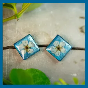 Zwei quadratische Ohrstecker aus blauem Epoxidharz, in denen jeweils eine kleine weiße getrocknete Blüte eingelassen ist.